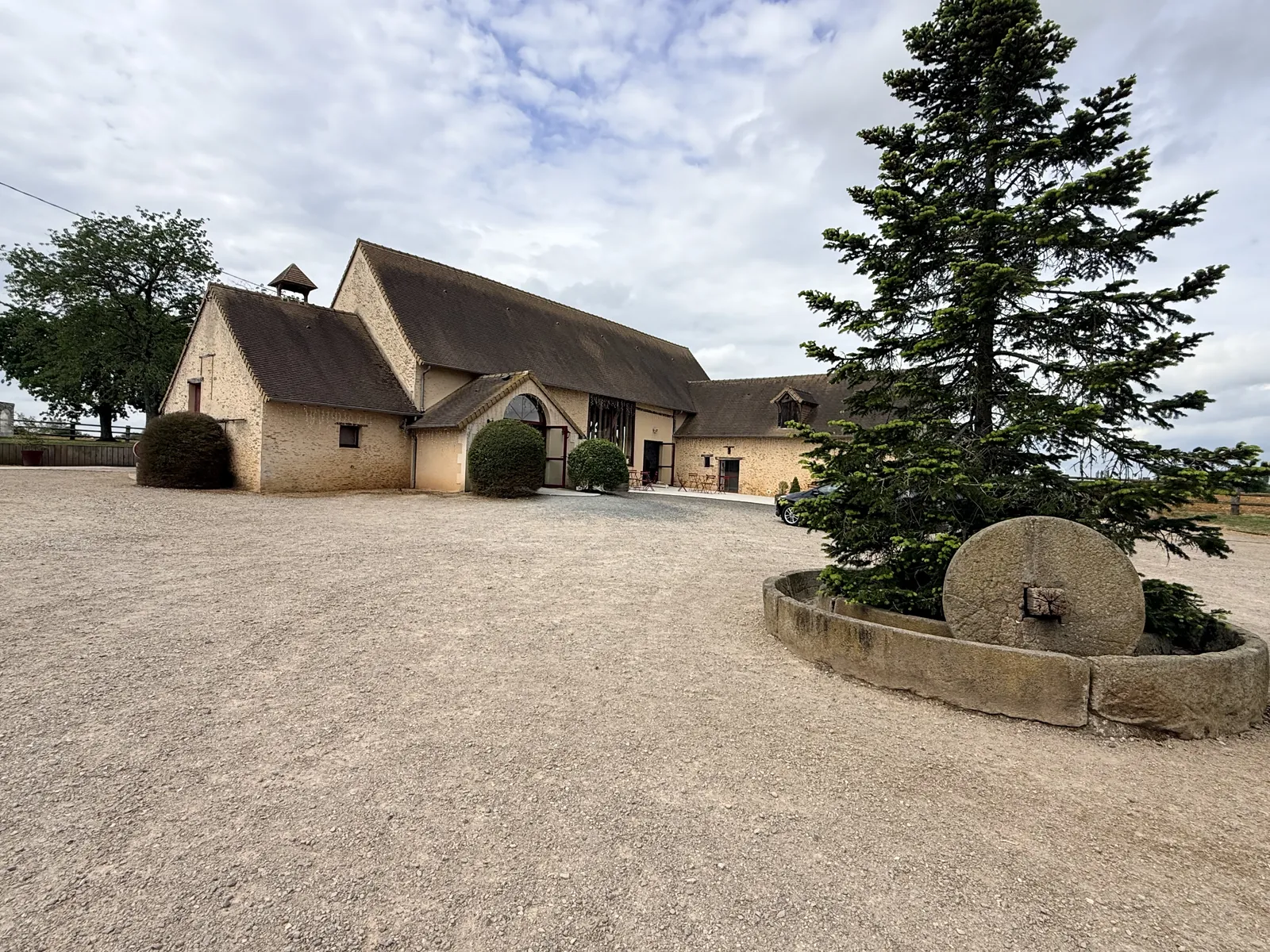 La cour gravillonnée, son sapin et sa meule de pierre devant la salle de réception